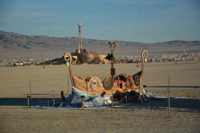The ship seen at distance across the open playa in daylight, dwarfed by the desert, with the Man and the mountains behind it