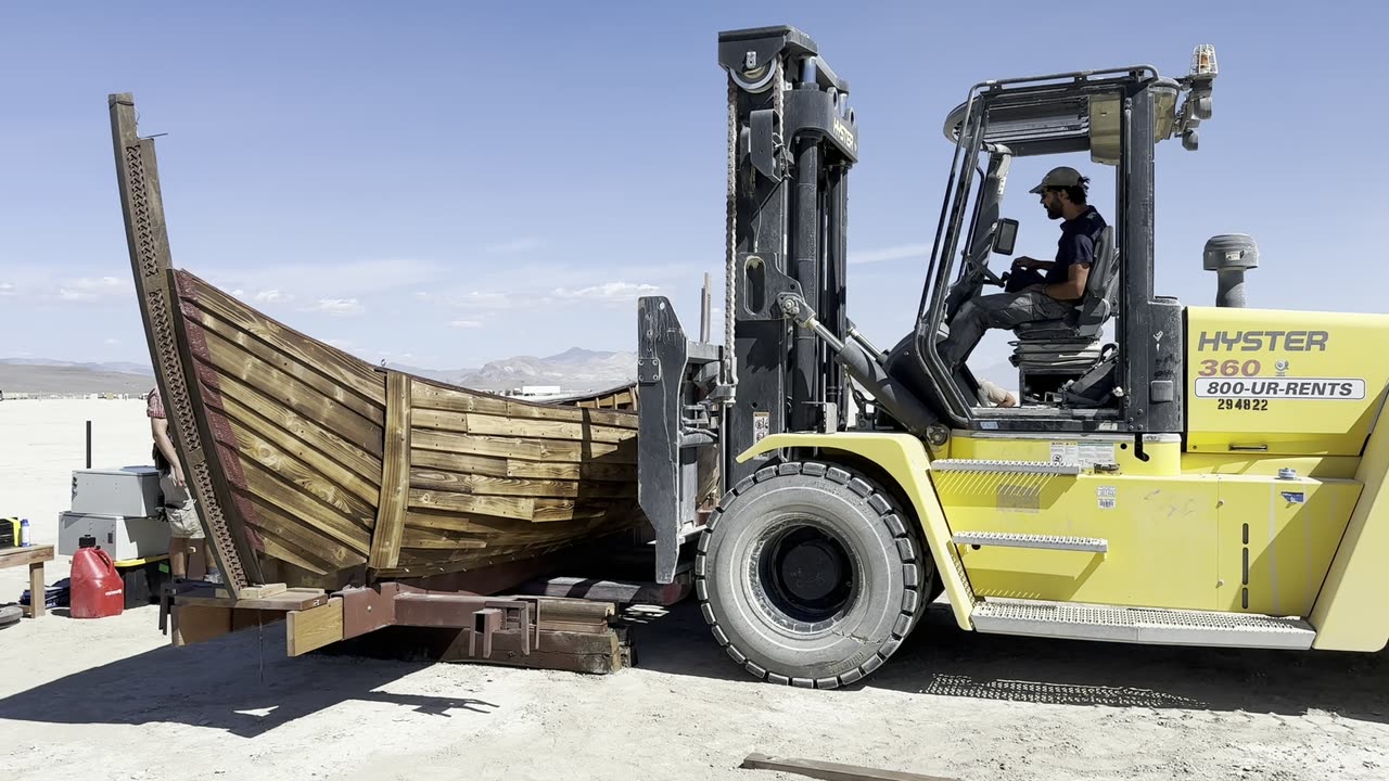 A forklift lowering a section of the ship's hull into place on the open playa