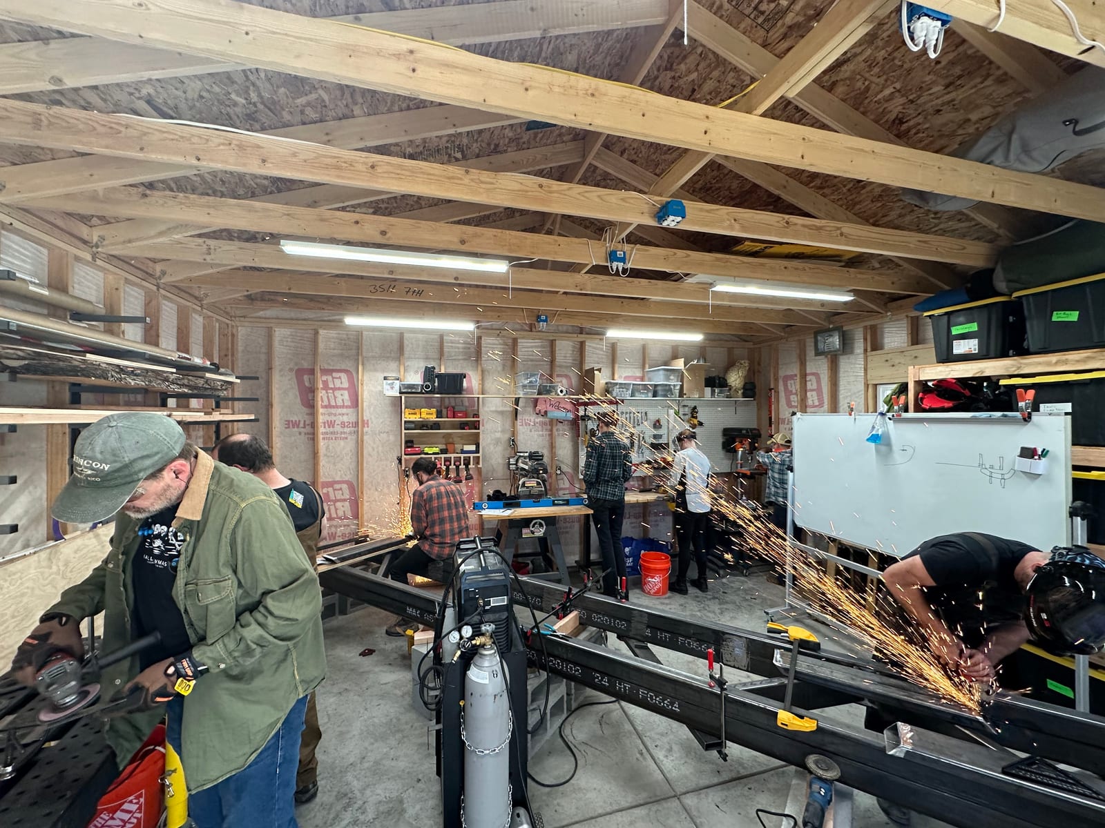 Build day inside a timber-framed shop, half a dozen people working along a run of steel box section while a grinder throws a sheet of sparks