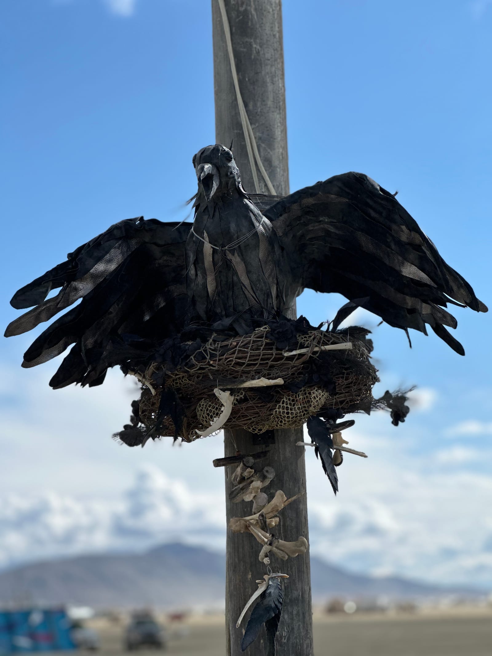 A life-size raven sculpture of black feathers and leather perched on a nest lashed to the mast, wings spread, against a blue desert sky
