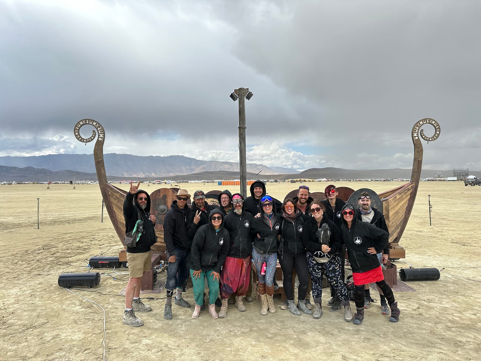 Sixteen crew members in matching black hoodies posed in front of the ship on the open playa under heavy gray cloud
