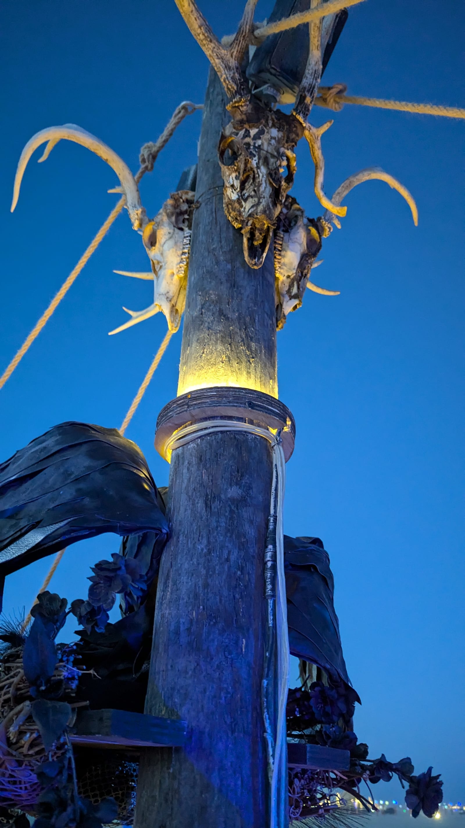 An antlered skull mounted high on the mast at blue hour, uplit from a collar of warm light at its base