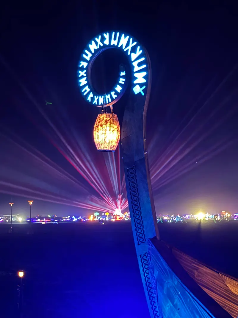 The ship's prow at night, a ring of illuminated blue runes above a glowing lantern, with laser beams from the city fanning out behind