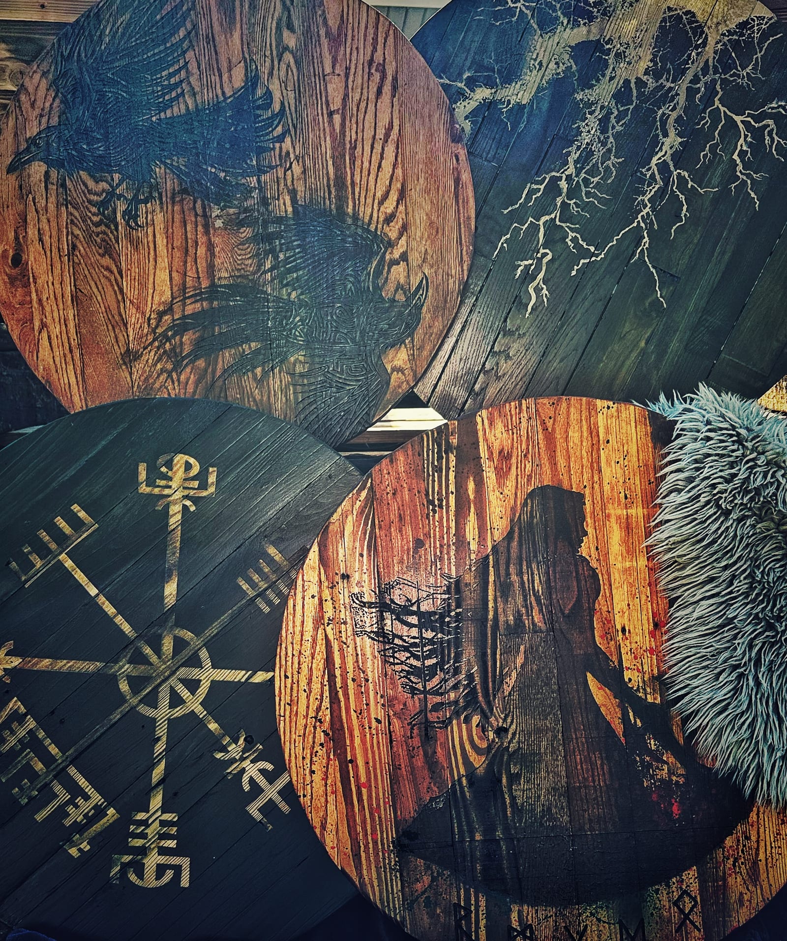 Four painted shields overlapping — a raven in flight on scorched pine, pale roots spreading across dark boards, a gold vegvísir stave, and a hooded figure in silhouette