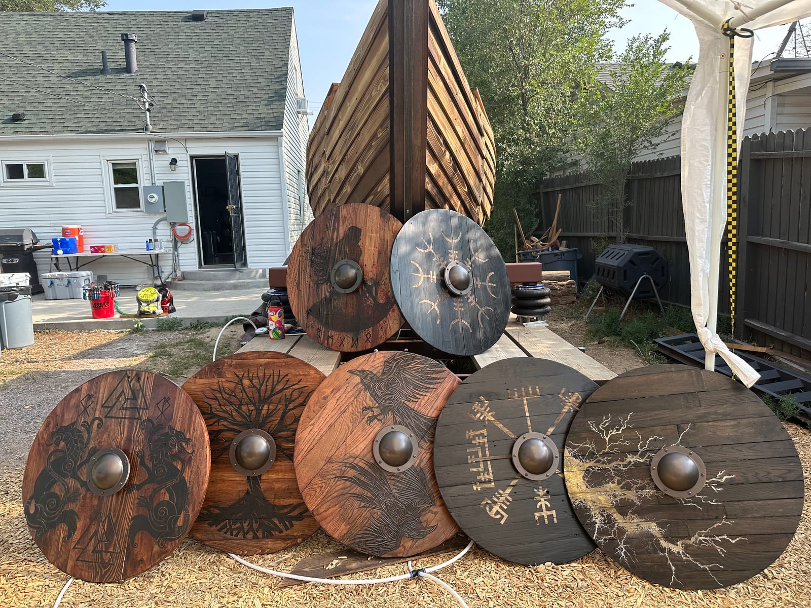 Seven painted round shields propped against the ship's hull in a suburban back yard, each with a different burned and stencilled design