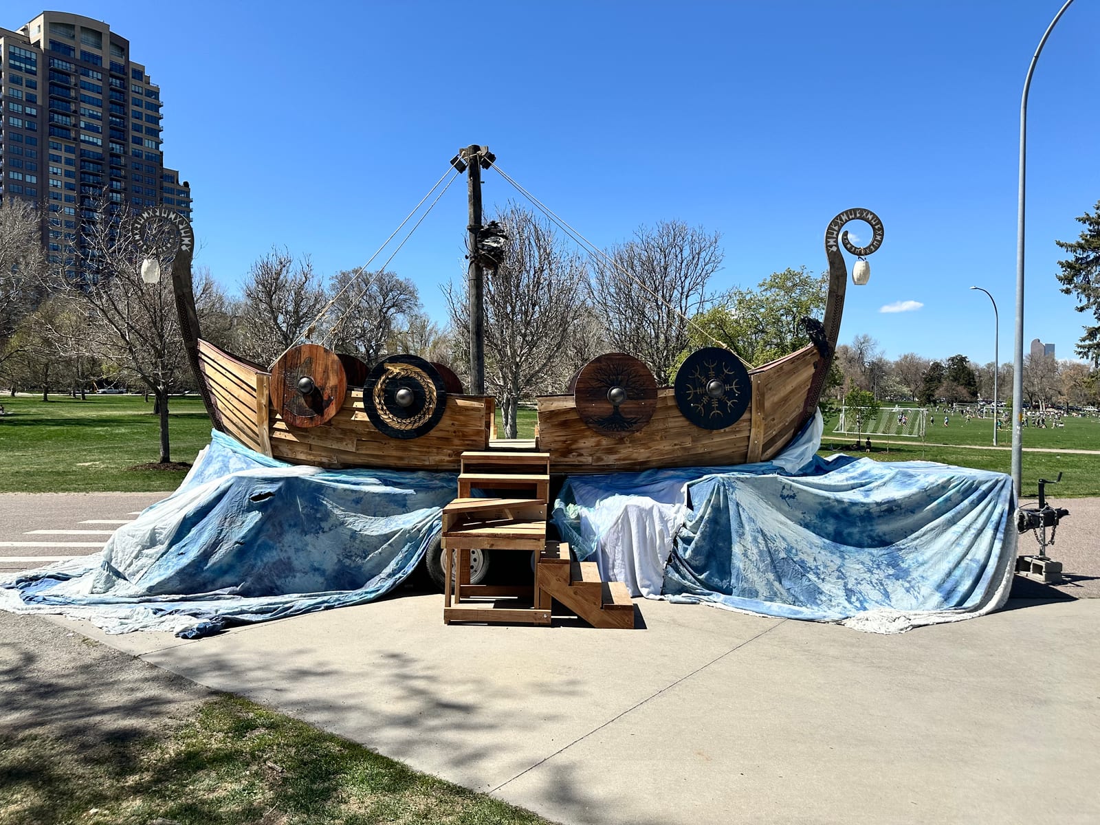 The complete ship parked on a path in a Denver park in spring, painted blue wave skirts spread around its base and wooden steps at the center