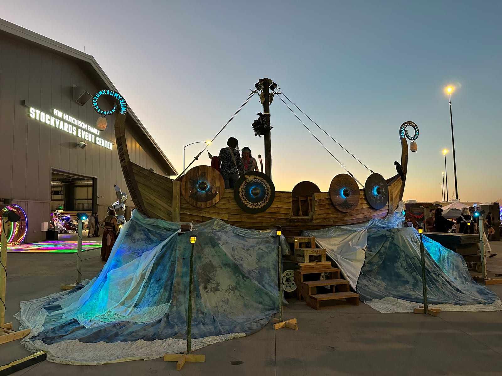 The ship lit at dusk outside an event center, shields glowing cyan and the wave skirts washed blue, people standing on deck