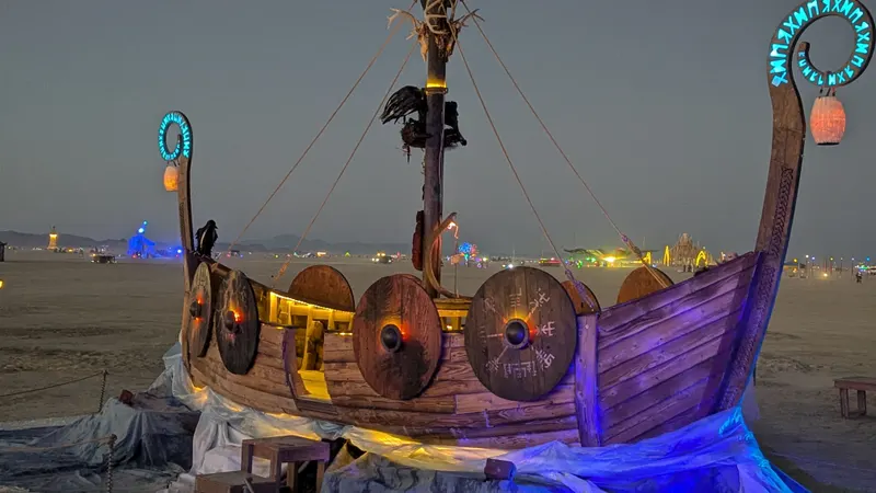 The finished viking ship at dusk on the playa, shields glowing warm from within and blue light washing the painted wave skirts at its base